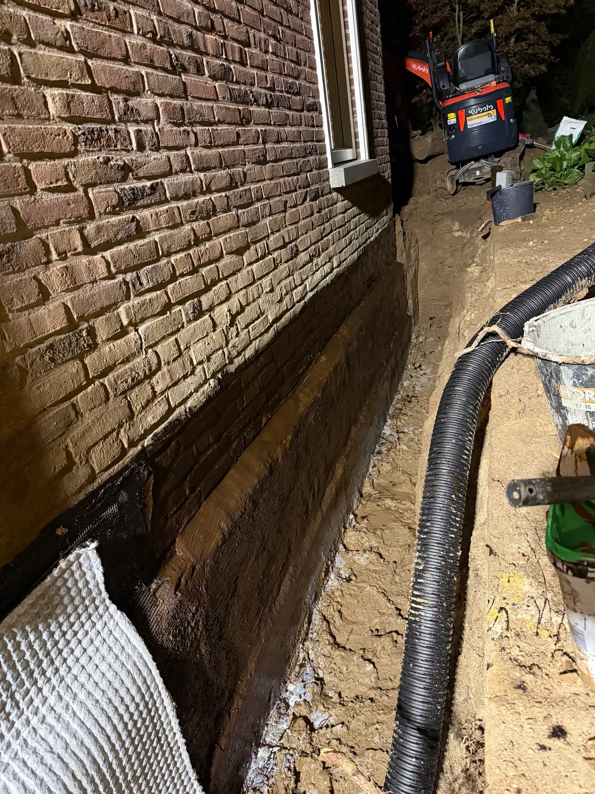 Bakstenen kelderwand met onder het maaiveld een zwarte bitumen dikcoating met wapeningsgaas, witte noppenfolie ertegen en een zwarte drainagebuis langs de sleuf, in Voerendaal