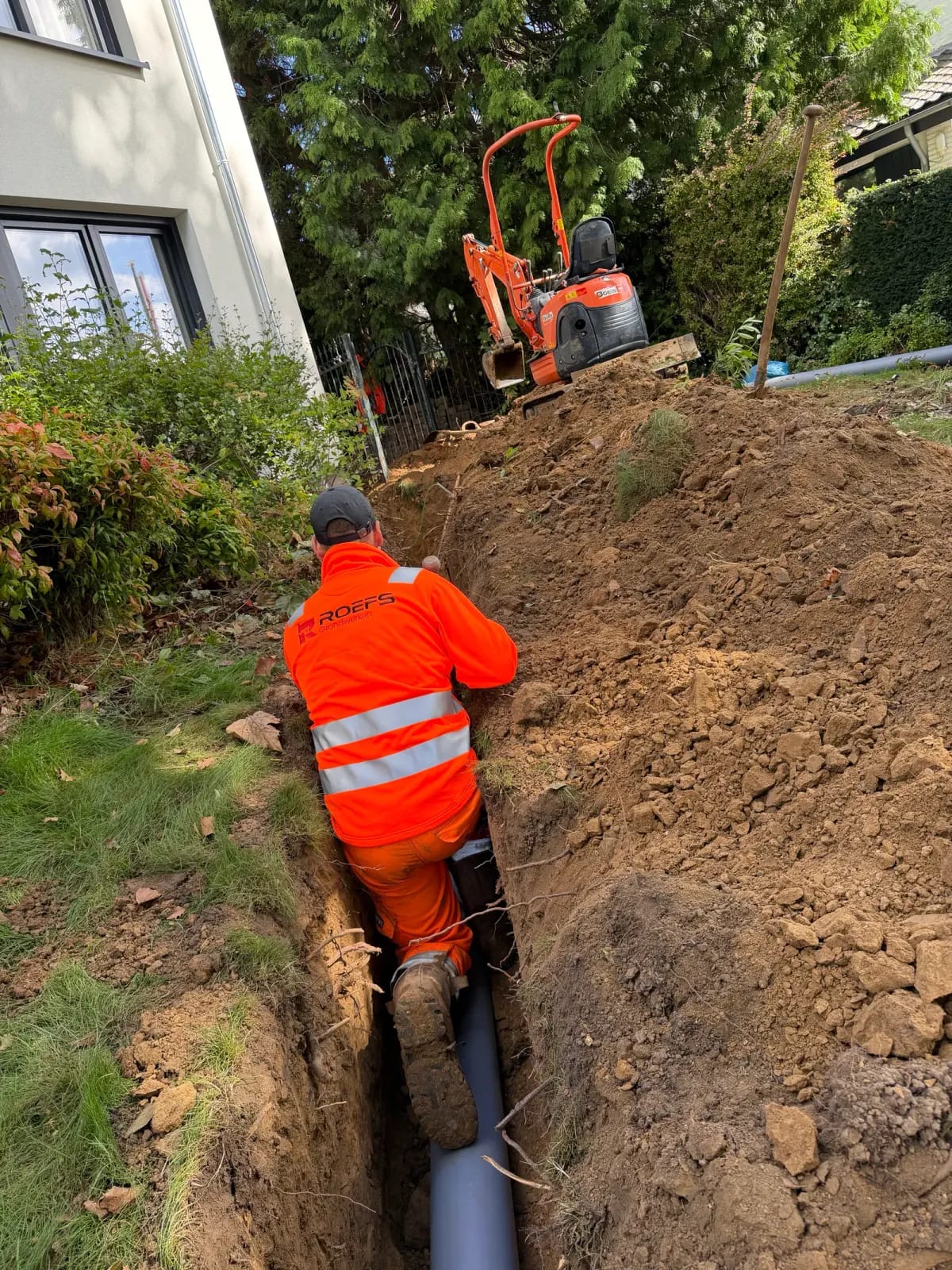 Dante Hoenjet in oranje werkkleding legt een rioolbuis in een smalle sleuf langs een woning in Meerssen, met een minigraver op de achtergrond