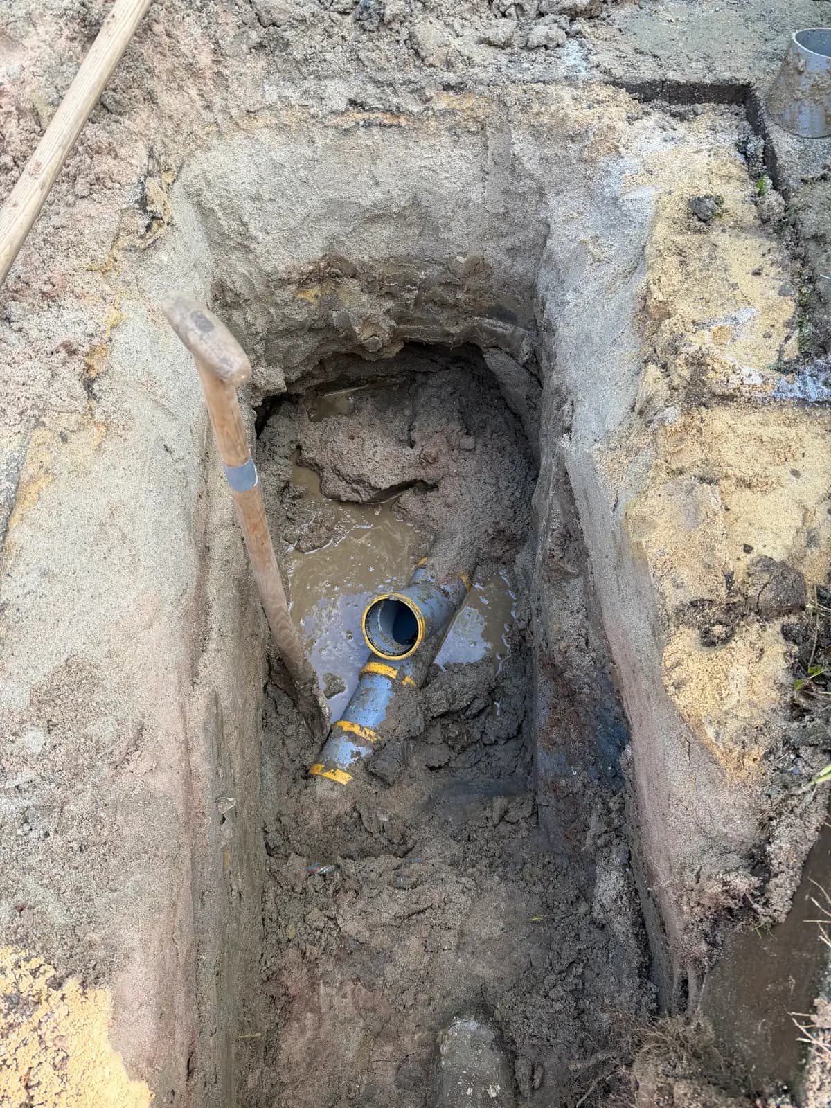 Uitgegraven put met grondwater op de bodem en een grijze pvc-leiding met een aftakking en gele ringen, bij de aansluiting op de bestaande riolering, in Voerendaal