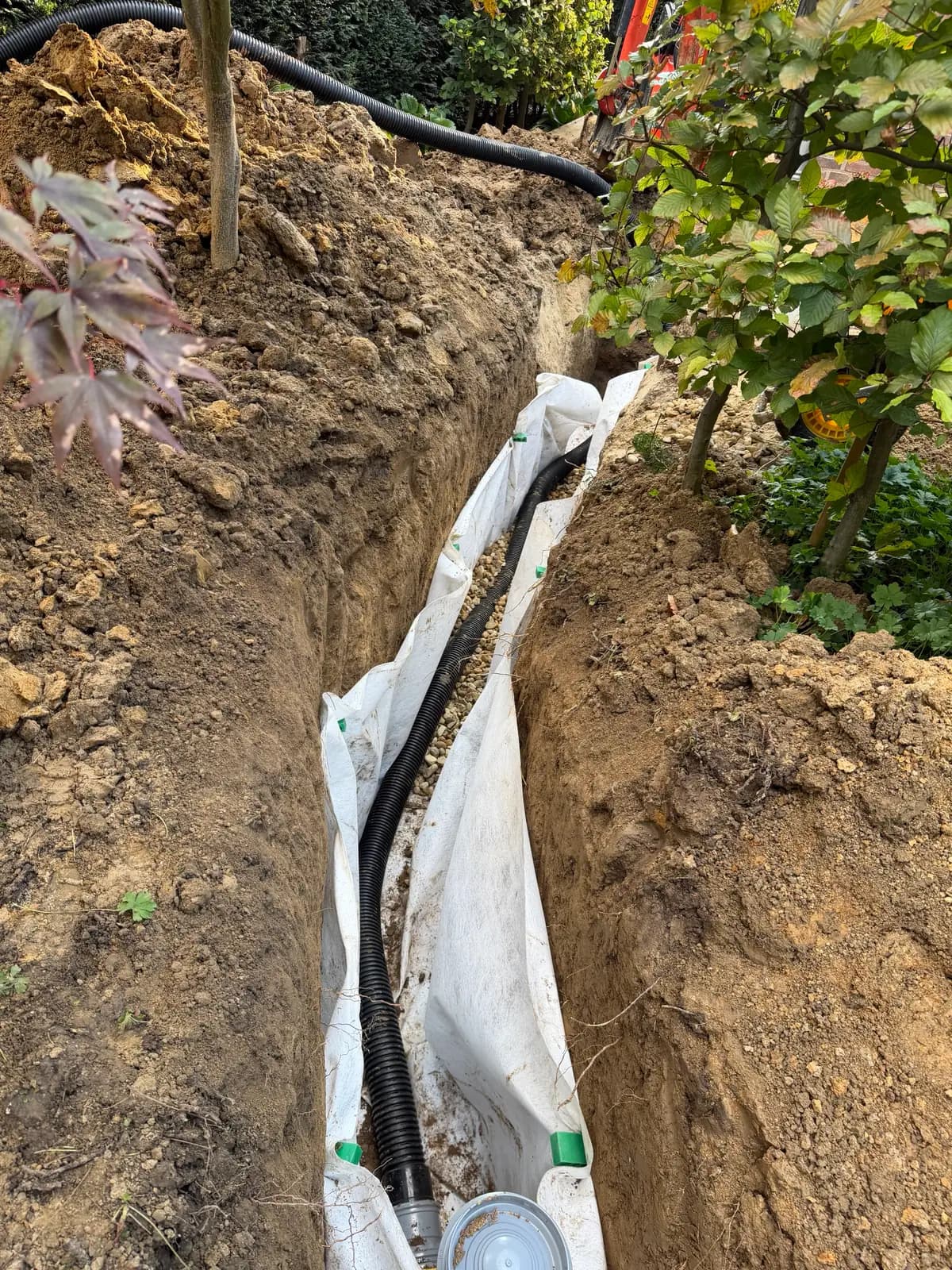 Sleuf met wit filterdoek langs de wanden, een bed van grind en een zwarte geribbelde drainagebuis die naar een grijze put met deksel loopt, in Voerendaal