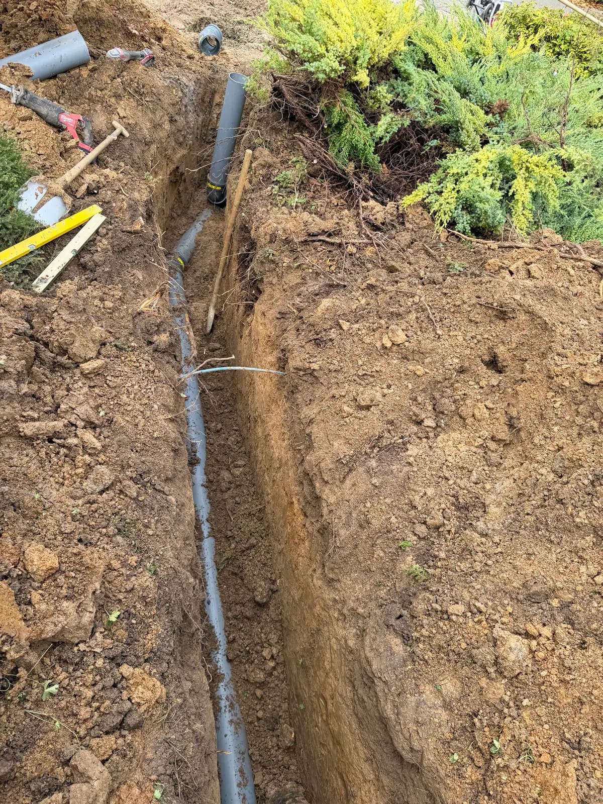 Nieuwe grijze pvc-rioolleiding in een smalle sleuf tussen coniferen, met hulpstukken, een waterpas en gereedschap naast de sleuf, in Voerendaal
