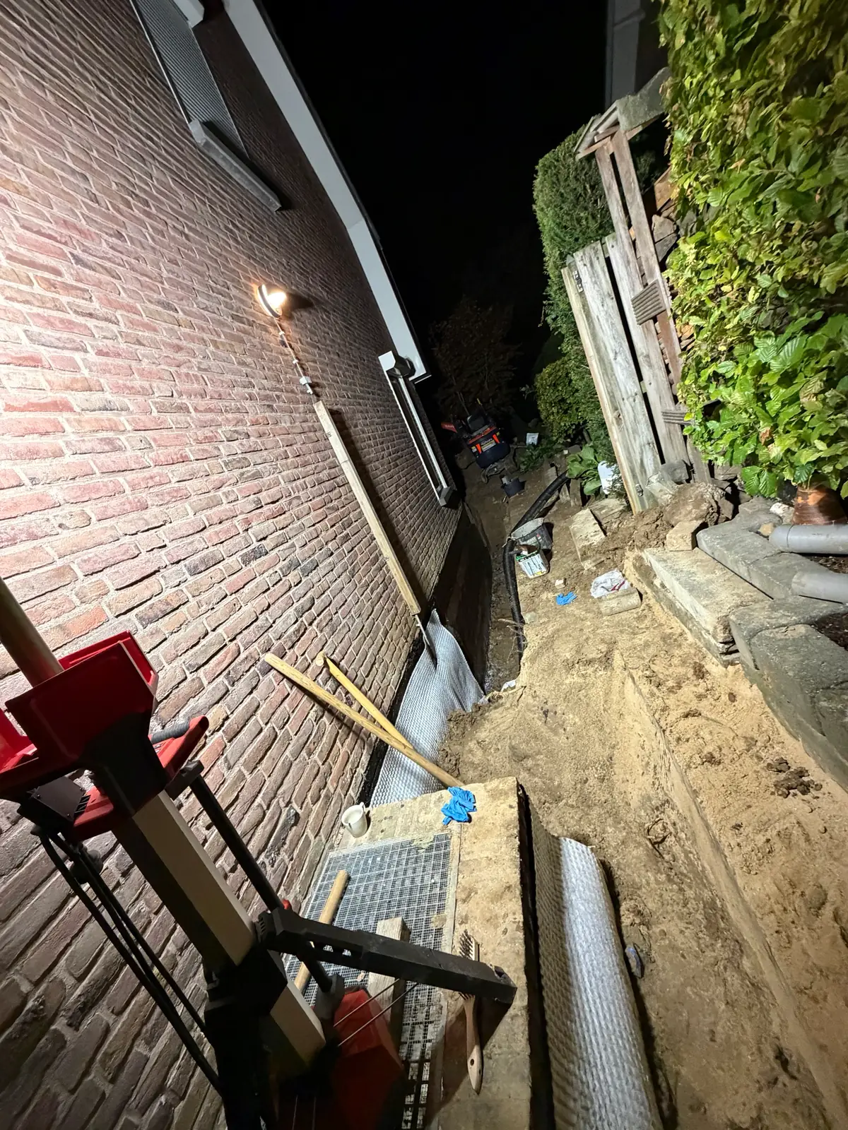 Sleuf langs de gevel van een bakstenen woning bij avond, met witte noppenfolie tegen de kelderwand, een rol noppenfolie en zand naast de sleuf, in Voerendaal